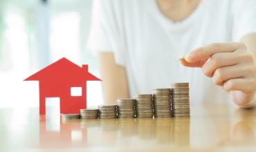 Down Payment Considered Top Barrier to Home Ownership Small red house with stacks of coins next to it on a counter with a person behind it