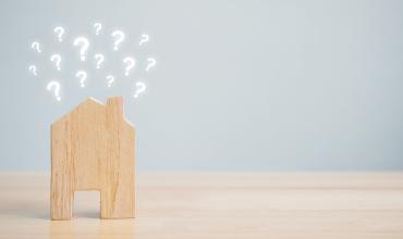 The Truth Behind 3 Common Home Buying Myths Small wooden house sitting on a desk with little question marks coming out of the roof