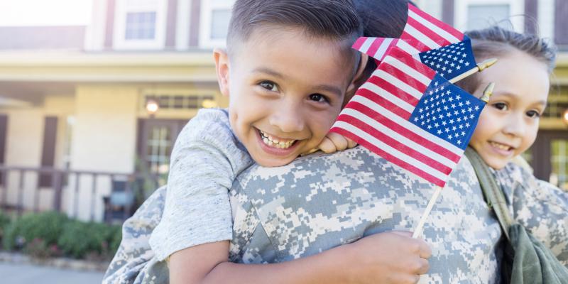 A child holding the US flag being held by their parent in military uniform 